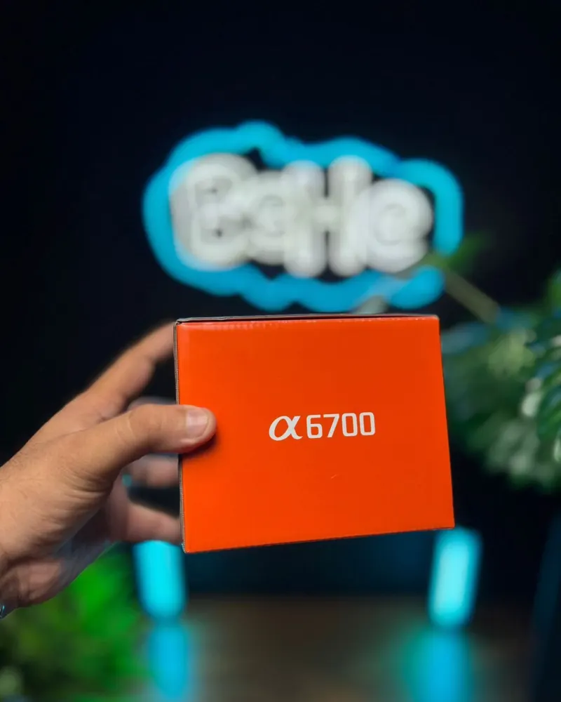 Caixa laranja da câmera Sony α6700 segurada na mão, em frente ao letreiro neon da BeHe